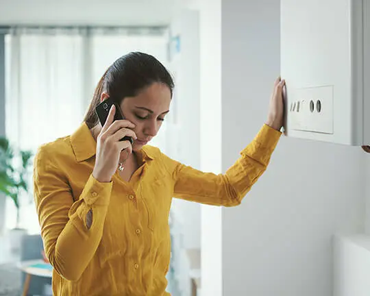 Femme au téléphone, l'air préoccupé, appuyée sur une chaudière murale blanche à l'intérieur d'une maison déclare une urgence plomberie